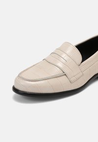 Mocassino in pelle beige con motivo coccodrillo, suola nera e due dettagli orizzontali sulla parte superiore, caratterizzato da una texture liscia e un design elegante.