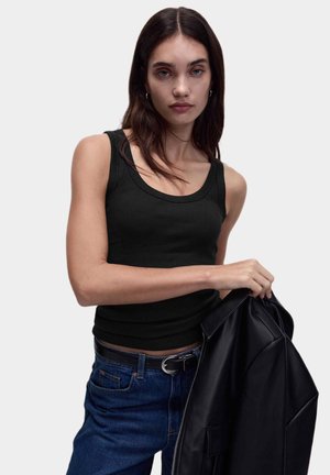 Schwarzes geripptes Tanktop mit rundem Halsausschnitt. Das Model hält eine schwarze Lederjacke und trägt dazu eine blaue Jeans und einen schwarzen Gürtel.