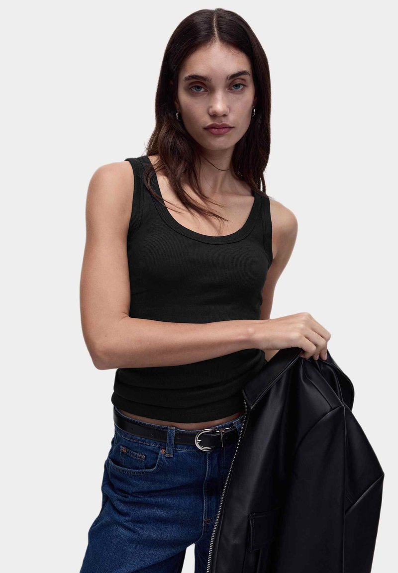 Débardeur noir à côtes avec un col rond. Le modèle tient une veste en cuir noire, associée à un jean en denim bleu et à une ceinture noire.
