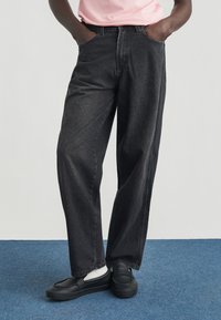 Μαύρο τζιν παντελόνι με άνετη εφαρμογή, μπροστινές τσέπες και διακοσμητικές ραφές, συνδυασμένο με μαύρα παπούτσια χωρίς κορδόνια και λευκές κάλτσες.