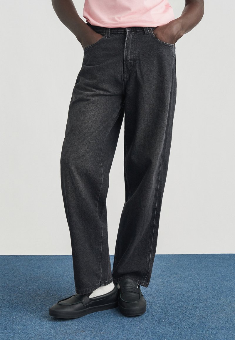 Zwarte spijkerbroek met een relaxte pasvorm, voorzakken en gestikte details, gecombineerd met zwarte instapschoenen en witte sokken.