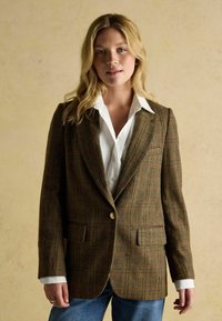 Pruun ruuduline bleiser, millel on nööbikinnitus, lõhega krae ja kaks esikot. Kantud valge kraega särgi kohal, koos siniste teksadega.