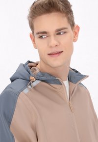 Jachetă cu glugă în bej și gri, cu fermoar frontal și șnur ajustabil. Fabricată dintr-un material ușor, cu o textură netedă.