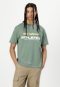 Zaļš kokvilnas T-krekls ar uzrakstiem "new balance" un "ATHLETICS" dzeltenā un baltā krāsā, ar mazu logo kreisajā pusē.