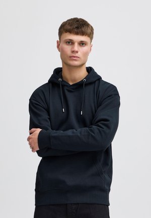 Jeune homme aux cheveux bruns courts portant un sweat à capuche noir, debout les bras croisés devant un fond gris clair uni.