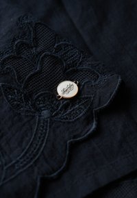 Marineblauwe stof met geborduurd bloemensysteem; heeft een ronde gouden metalen tag met het woord "Superdry" erop, bevestigd aan de textiel.