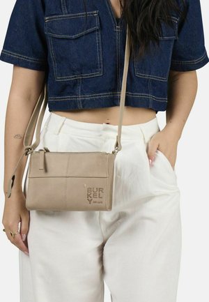 Borsa a tracolla in pelle beige con chiusura a zip e stampa del logo. Presenta una tasca frontale piatta e una tracolla regolabile. Indossata con una blusa in denim e pantaloni bianchi.