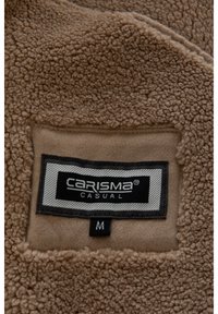 Pruun fliiskangas tekstuurse pinnaga. Silt, millel on kiri „CARISMA CASUAL” ja suurus „M”, on paigutatud siledale alusele musta ja halli toonidega aktsentidega.