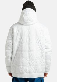 Biała pikowana kurtka puchowa z kapturem, z teksturowanym geometrycznym wzorem i elastycznymi mankietami, noszona z czarną dzianinową czapką.
