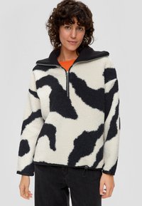 Polarowy półgolf w odcieniu off-white z czarnym nadrukiem w krowie plamy, z zamek przy kołnierzu, długimi rękawami i lekko ściągniętym dołem.