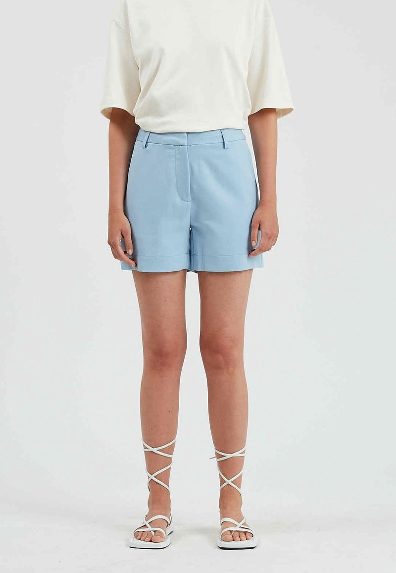Shorts bleu clair ajustés avec une coupe droite, poches latérales et texture lisse. Associés à un haut blanc à manches courtes et des sandales blanches.