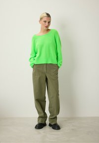 Neon žalias megztinis su laisvu siluetu, derinamas su laisvomis tamsiai žaliomis kelnėmis. Juodi masyvūs batai užbaigia aprangą prieš neutralų foną.