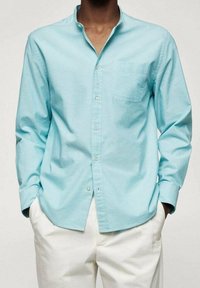 Hellblaues Langarmhemd aus Baumwolle mit Button-Down-Kragen und Brusttasche. Getragen über einer weißen Hose.