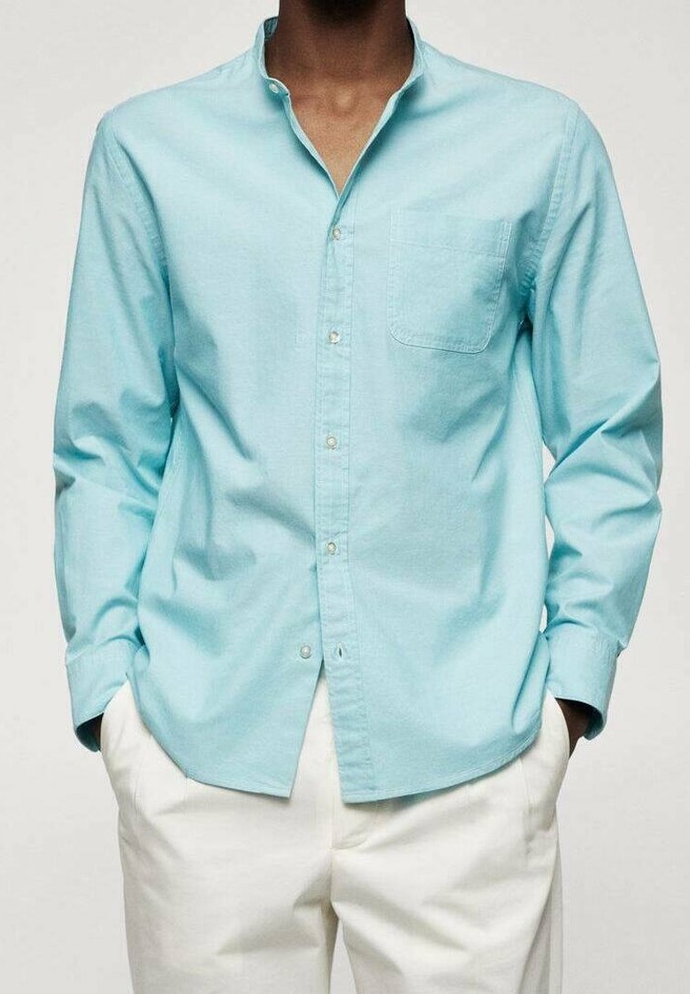 Hellblaues Langarmhemd aus Baumwolle mit Button-Down-Kragen und Brusttasche. Getragen über einer weißen Hose.