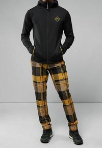 Felpa nera con zip e logo, abbinata a pantaloni a quadri nei toni giallo, marrone e nero. Il tessuto testurizzato e le sneakers sportive completano l'outfit.