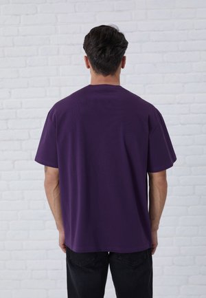 Fioletowa bawełniana koszulka z krótkim rękawem i luźnym krojem. Bezszwowy dekolt i gładka struktura. Na sobie czarne dżinsy.