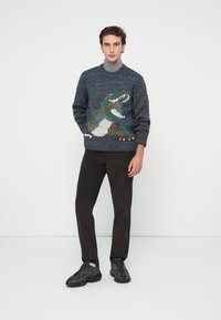 Un pull en tricot bleu et vert, arborant un motif de dinosaure texturé. Associé à un pantalon en velours côtelé noir et des chaussures de sport noires.