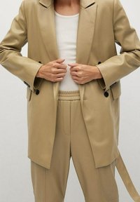 Beige op maat gemaakt blazer met scherpe revers, dubbele knopen en een ontspannen pasvorm, gecombineerd met bijpassende afhangende broeken met een elastische tailleband.