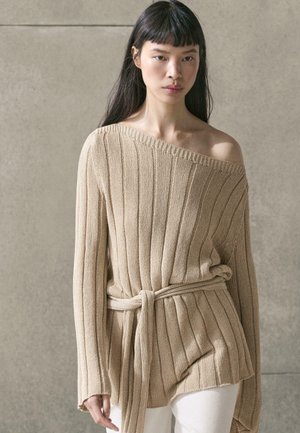 Vrouw met lang donker haar draagt een beige geribbelde gebreide trui met een off-shoulder halslijn en een riem, gecombineerd met witte broek, neutrale achtergrond.