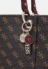 Zwarte gestructureerde totebag met een bruin logo patroon, voorzien van bordeauxkleurige handvatten en gouden hardware, inclusief sleutelhanger-achtige logo charme.