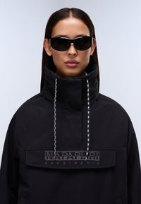 Mujer con cabello largo y oscuro, usando gafas de sol negras y una chaqueta negra de cuello alto con la marca "Napapijri geographic".