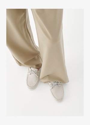 Mocassins en daim beige avec lacets portés avec un pantalon ample marron clair sur un fond blanc uni.