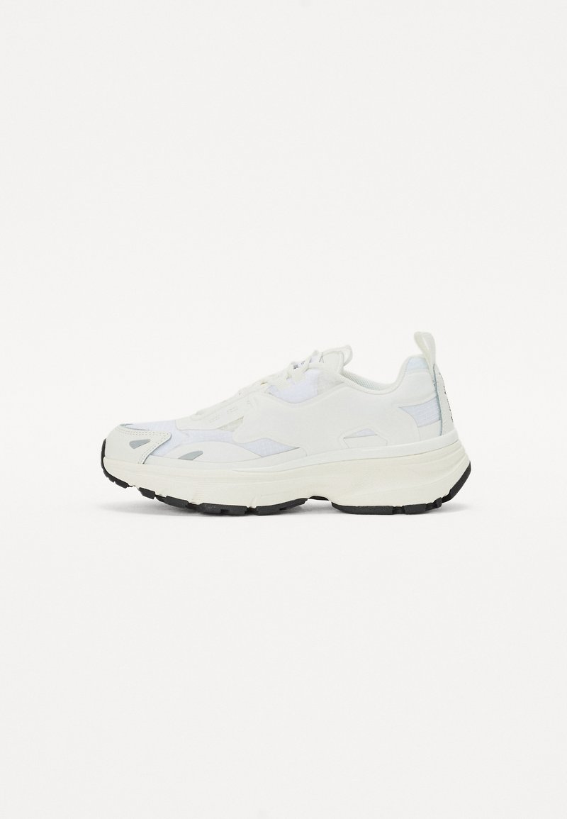 Fehér sportcipő hálós és szintetikus anyagból, vastag talppal, kerek orral, fekete talppal és diszkrét dizájn részletekkel a felső részen.