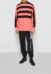 Růžová a černá pruhovaná košile s dlouhým rukávem a černým límečkem s nápisem "Moschino" v žluté barvě, doplněná černými kalhotami s bílým potiskem loga.