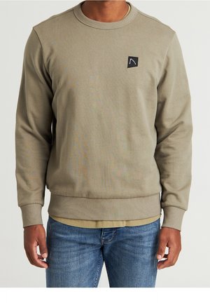 Homme portant un sweat-shirt kaki à col ras du cou avec un petit patch logo noir et blanc sur la poitrine, associé à un jean bleu.