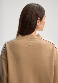 Felpa beige con collo alto, caratterizzata da strisce laterali multicolori e un logo metallico sul retro. Texture liscia.