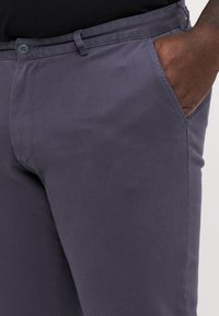 Graue Baumwollhosen mit glatter Textur, ausgestattet mit Seitentaschen und einem Knopfverschluss. Sichtbare Nähte entlang der Säume für zusätzliche Details.