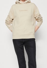 Felpa beige con cappuccio e tasca a marsupio, caratterizzata da un logo "Calvin Klein" tono su tono sul petto. Il tessuto appare morbido e liscio.