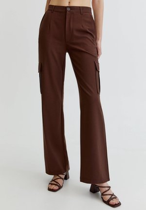 Kobieta w brązowych, prosto krojonych spodniach cargo z wysokim stanem, bocznymi kieszeniami oraz brązowych sandałach na słupku z paskami, na jednolitym tle.