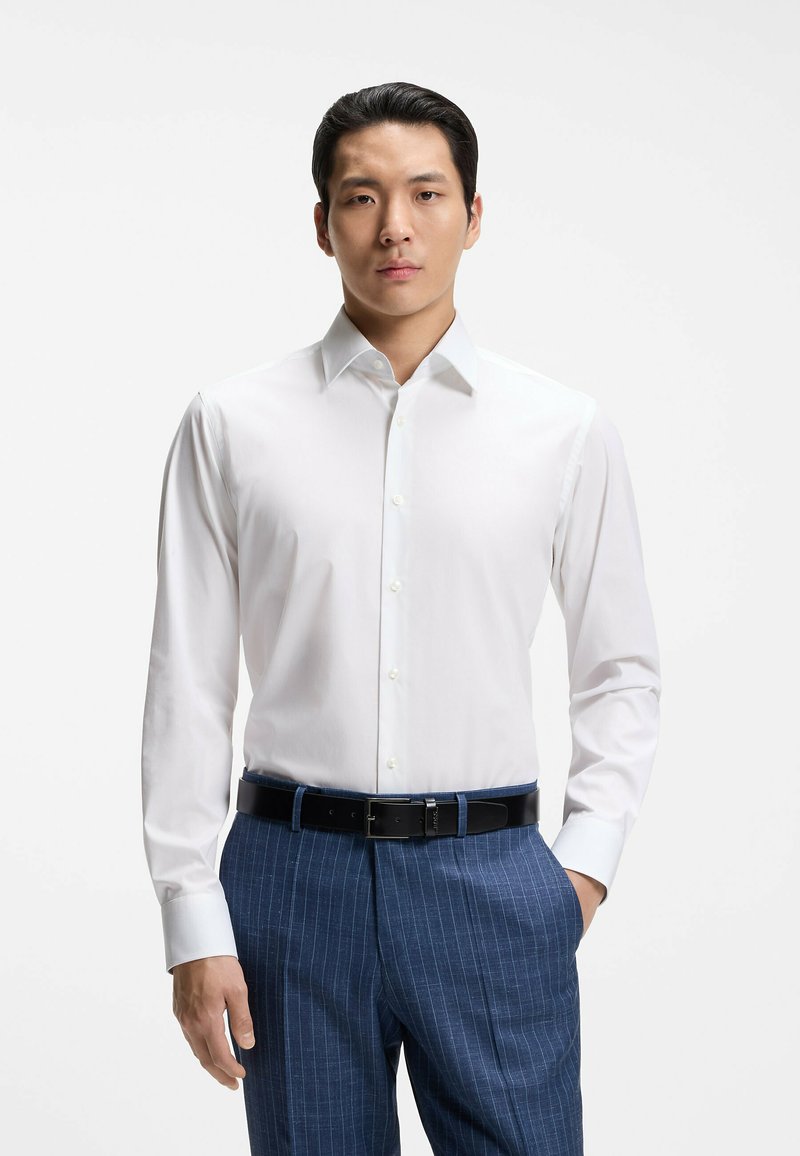 Homme portant une chemise blanche à manches longues, un pantalon bleu à fines rayures et une ceinture noire, debout avec une main dans la poche.