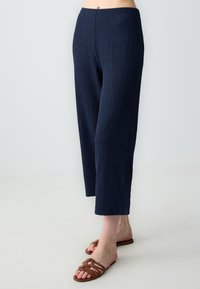 Pantalons taille large texturés bleu marine avec un design froissé. Arrivant à la cheville, assortis à des sandales à lanières marron.