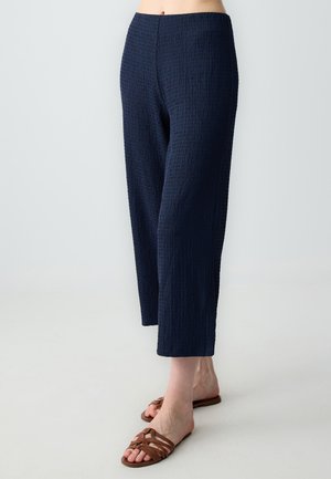 Pantaloni a gamba larga blu navy con una texture crepata. Si fermano alla caviglia, abbinati a sandali marroni con fasce.