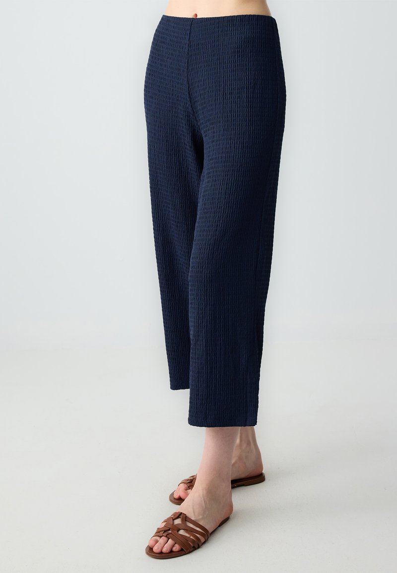 Pantalons taille large texturés bleu marine avec un design froissé. Arrivant à la cheville, assortis à des sandales à lanières marron.