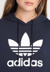 Marinblå huva med en vit Adidas-logotyp med bladdesign; tillverkad av strukturerad tyg med snörning och ribbad detaljer.