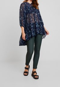 Námorně modrá průhledná blůza s květinovým vzorem, vysokým výstřihem a volánkovým lemem, v kombinaci s tmavě zelenými skinny džíny a černými sandály na tenkých páscích.