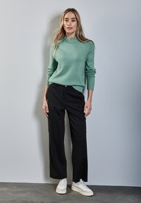 Mintgrüner Strickpullover mit geripptem Kragen, kombiniert mit locker sitzenden schwarzen Hosen und weißen Sneakers vor einem hellgrauen Hintergrund.