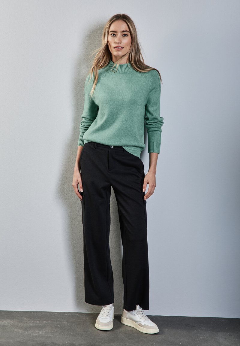 Mintgrüner Strickpullover mit geripptem Kragen, kombiniert mit locker sitzenden schwarzen Hosen und weißen Sneakers vor einem hellgrauen Hintergrund.