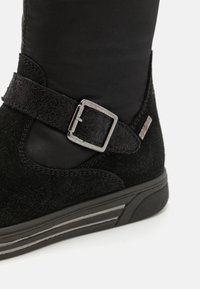 Bottine noire avec une finition en daim, une sangle texturée avec une boucle en argent, et une semelle en caoutchouc basse avec des rayures horizontales.