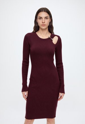 Femme portant une robe bordeaux, ajustée, côtelée, à manches longues, avec un détail de fermoir doré sur une découpe près de l'épaule droite.