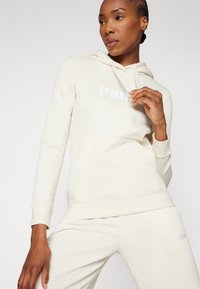 Krämfärgad hoodie tillverkad av mjukt tyg, med en framficka i kängurustil och en tryckt logotyp i vitt. Matchande krämfärgade byxor syns nedanför.