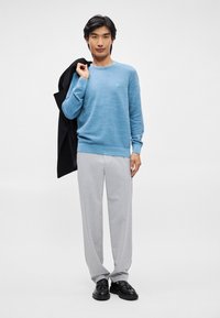 Μπλε πουλόβερ σε υφή πλέξης, συνδυασμένο με ανοιχτό γκρι παντελόνι. Μαύρο σακάκι περασμένο στον έναν ώμο. Μαύρα παπούτσια με υφή.