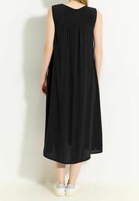 Svart ärmlös klänning med rundad halsringning, rynkad detalj i ryggen och lös passform, som når under knäna. Bärs med vita sneakers.