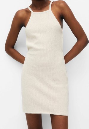 Robe en maille beige sans manches et ajustée avec fines bretelles, portée par une personne à la peau foncée, debout, les mains derrière le dos.
