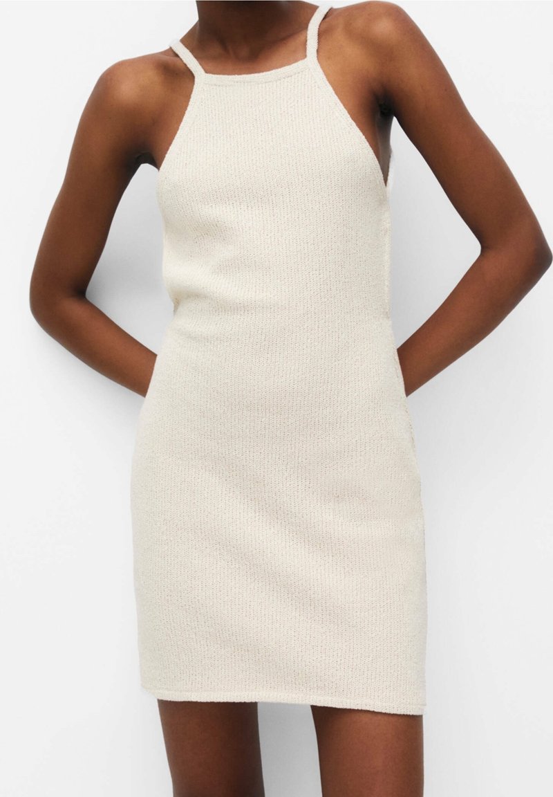 Robe en maille beige sans manches et ajustée avec fines bretelles, portée par une personne à la peau foncée, debout, les mains derrière le dos.