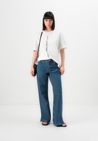 Λευκό oversized t-shirt με κάθετο μαύρο κείμενο, συνδυασμένο με φαρδιά τζιν παντελόνια μπλε. Μαύρη τσάντα χιαστί και επίπεδες μαύρες σανδάλες.