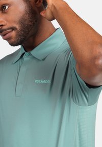 Světle tyrkysové polo tričko má klasický límec, knoflíkovou légu, logo Rossignol a strukturované boční panely pro prodyšnost.